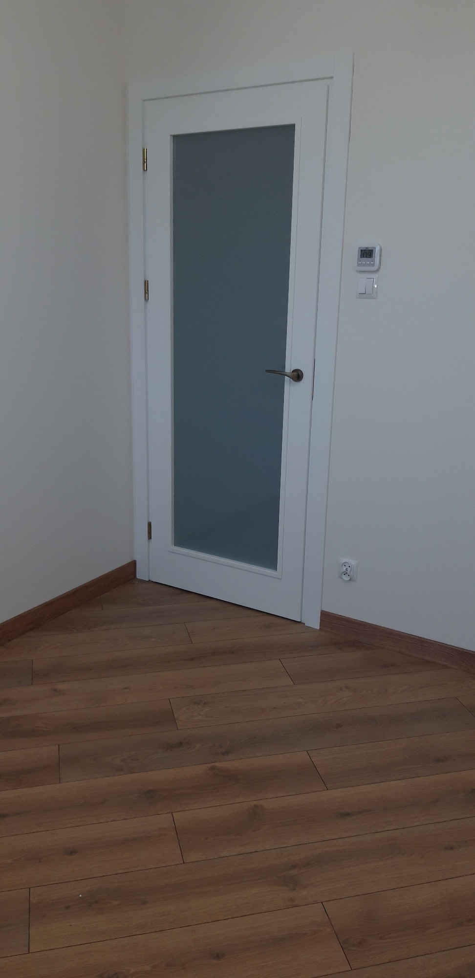 Białe drzwi wewnętrzne z matową szybą na tle drewnianej podłogi, widoczny termostat pokojowy i gniazdko elektryczne.