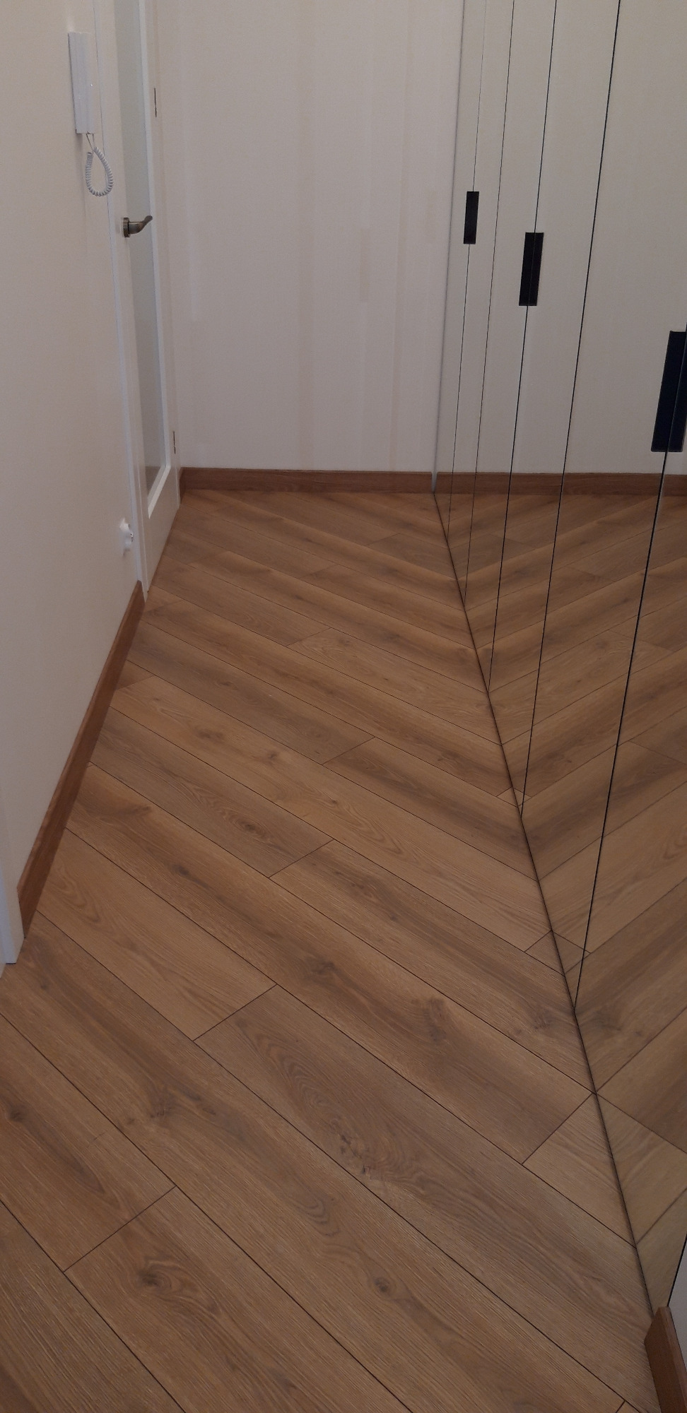 Ułożona w jodełkę podłoga z paneli drewnopodobnych, widok z perspektywy podłogi w kierunku drzwi i lustrzanej szafy.