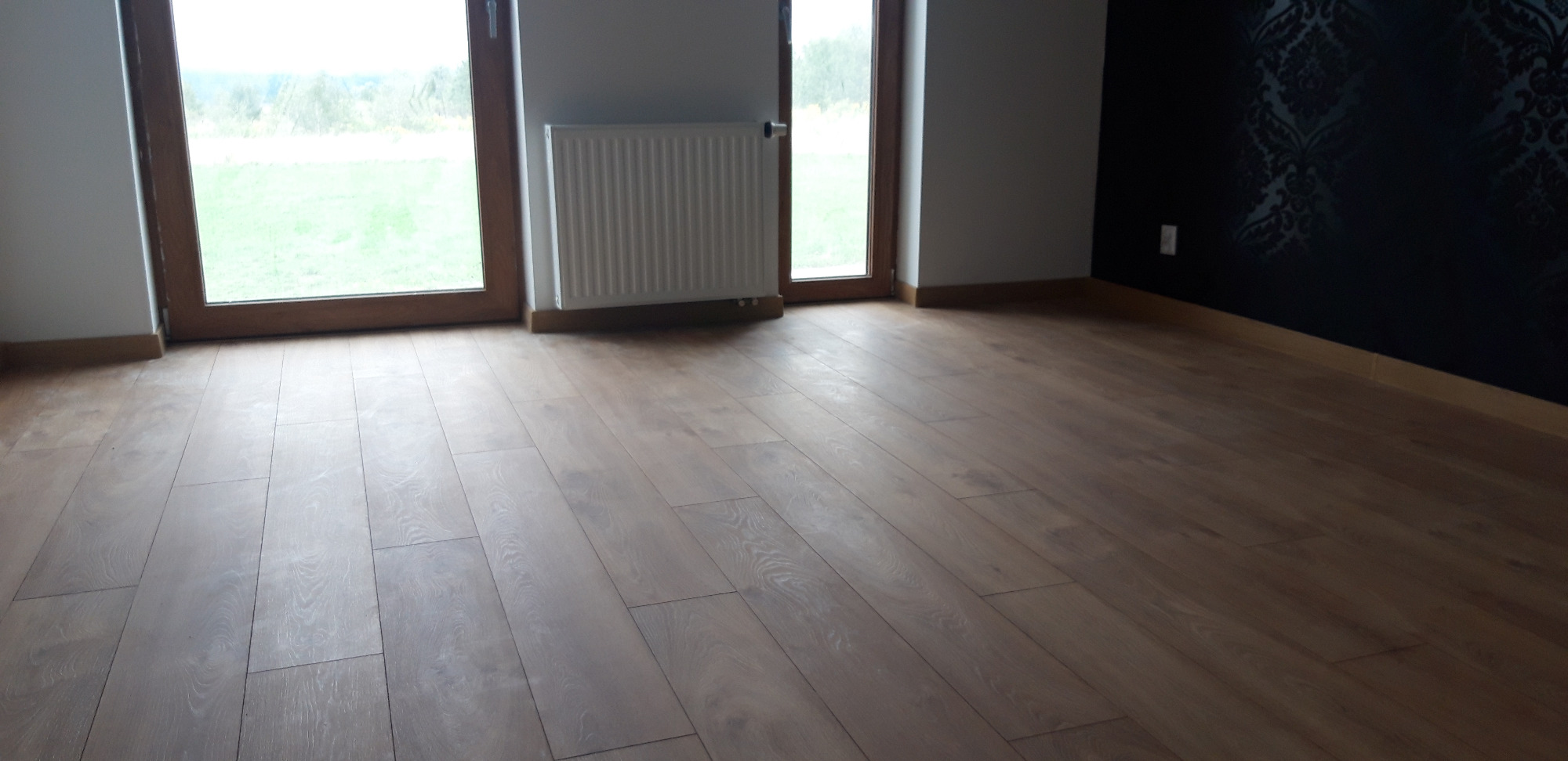 Ułożona jasnobrązowa podłoga z paneli, widok na okno z widokiem na zieleń oraz czarną ścianę z wzorem w tle, radiator pod oknem.