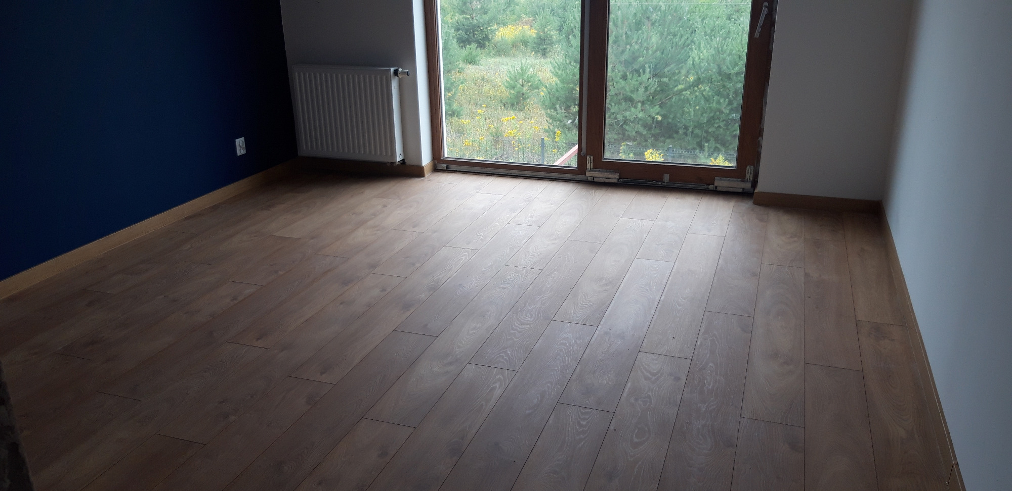 Jasne, drewnopodobne panele ułożone w nowym pokoju z widokiem na zieleń za oknem, niebieska ściana po lewej.