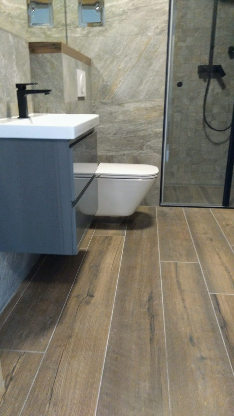 Nowoczesna łazienka z drewnopodobną podłogą, wiszącą toaletą i kabiną prysznicową z czarnymi elementami, minimalistyczna umywalka z czarną baterią.