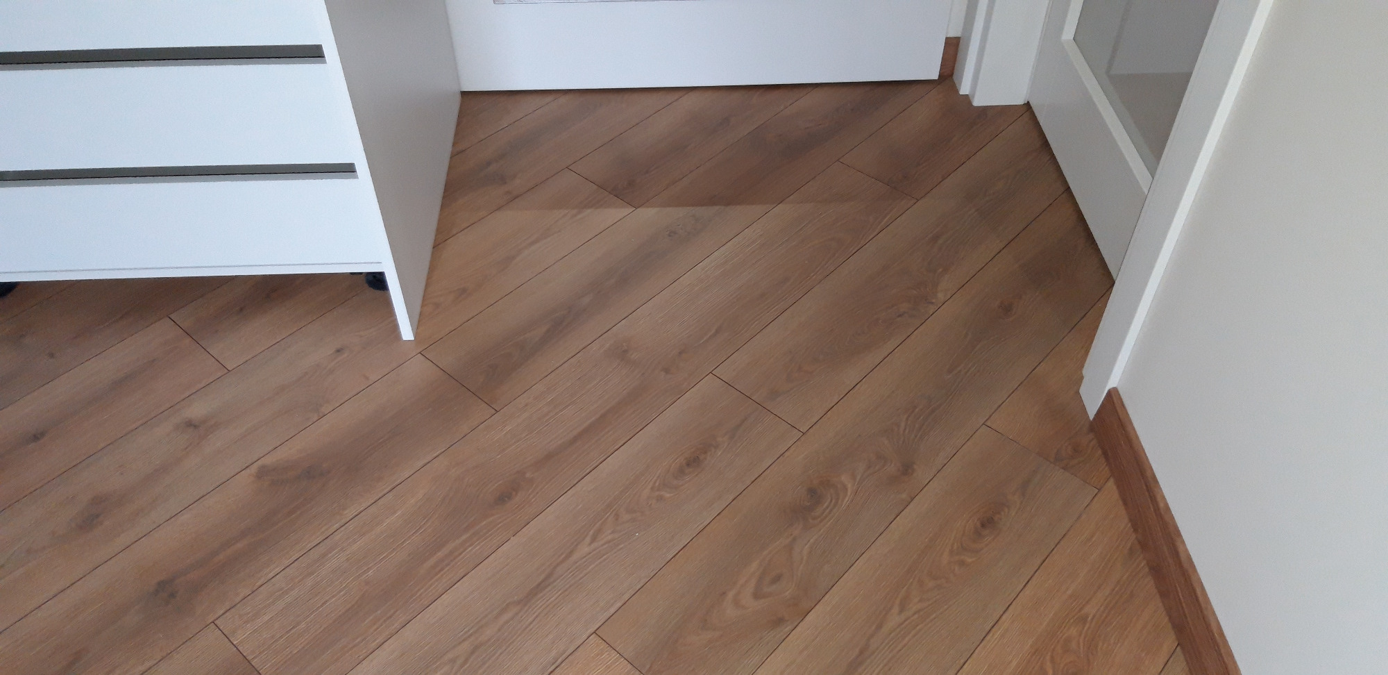 Ułożone panele podłogowe w jodełkę, widoczny narożnik białej szafki z szufladami na kółkach i fragment otwartych białych drzwi.