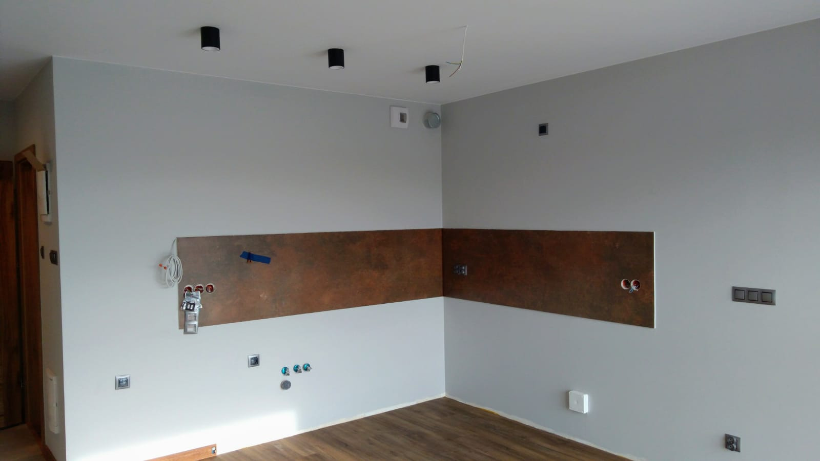 Wnętrze w trakcie remontu: szare ściany z poziomym pasem dekoracyjnym imitującym rdzę, widoczne puszki elektryczne, kable, gniazdka i włączniki w trakcie montażu, drewnopodobna podłoga.