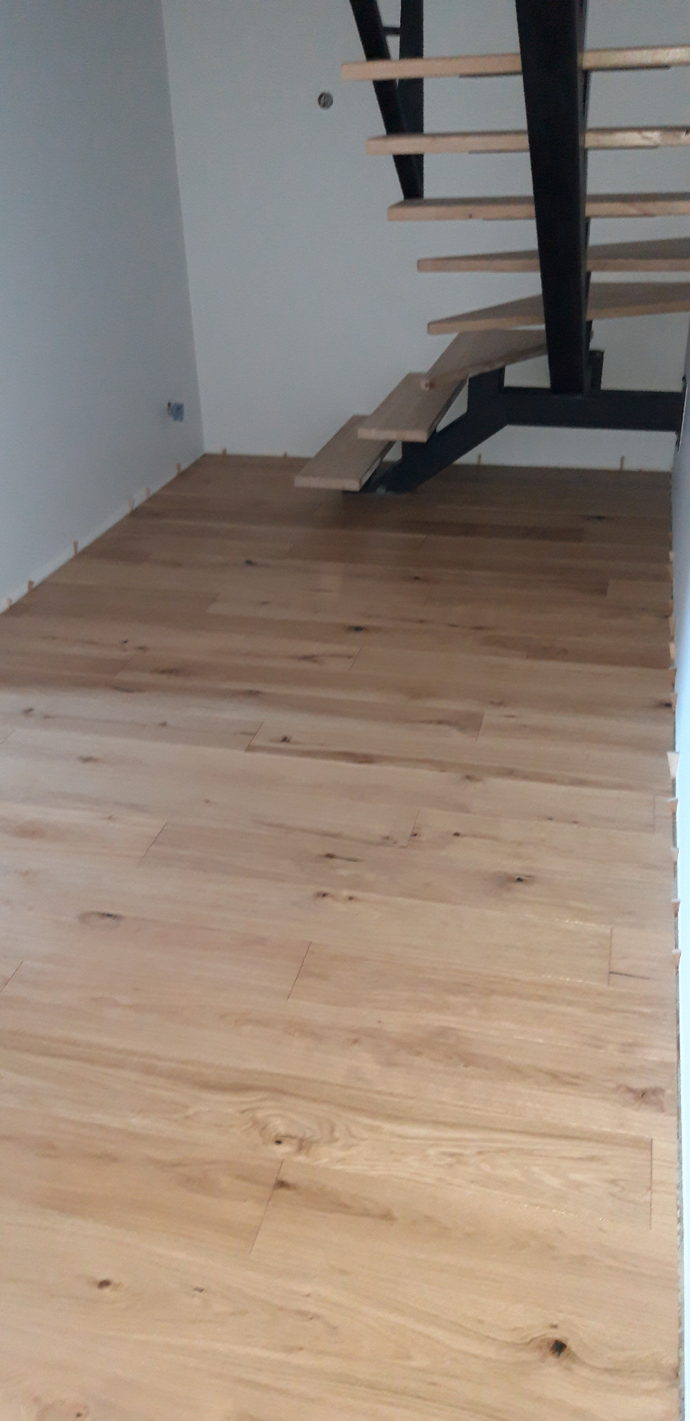 Jasne, drewniane deski podłogowe ułożone w pomieszczeniu z minimalistycznymi schodami na metalowej konstrukcji, widoczne kliny dystansowe przy ścianach.