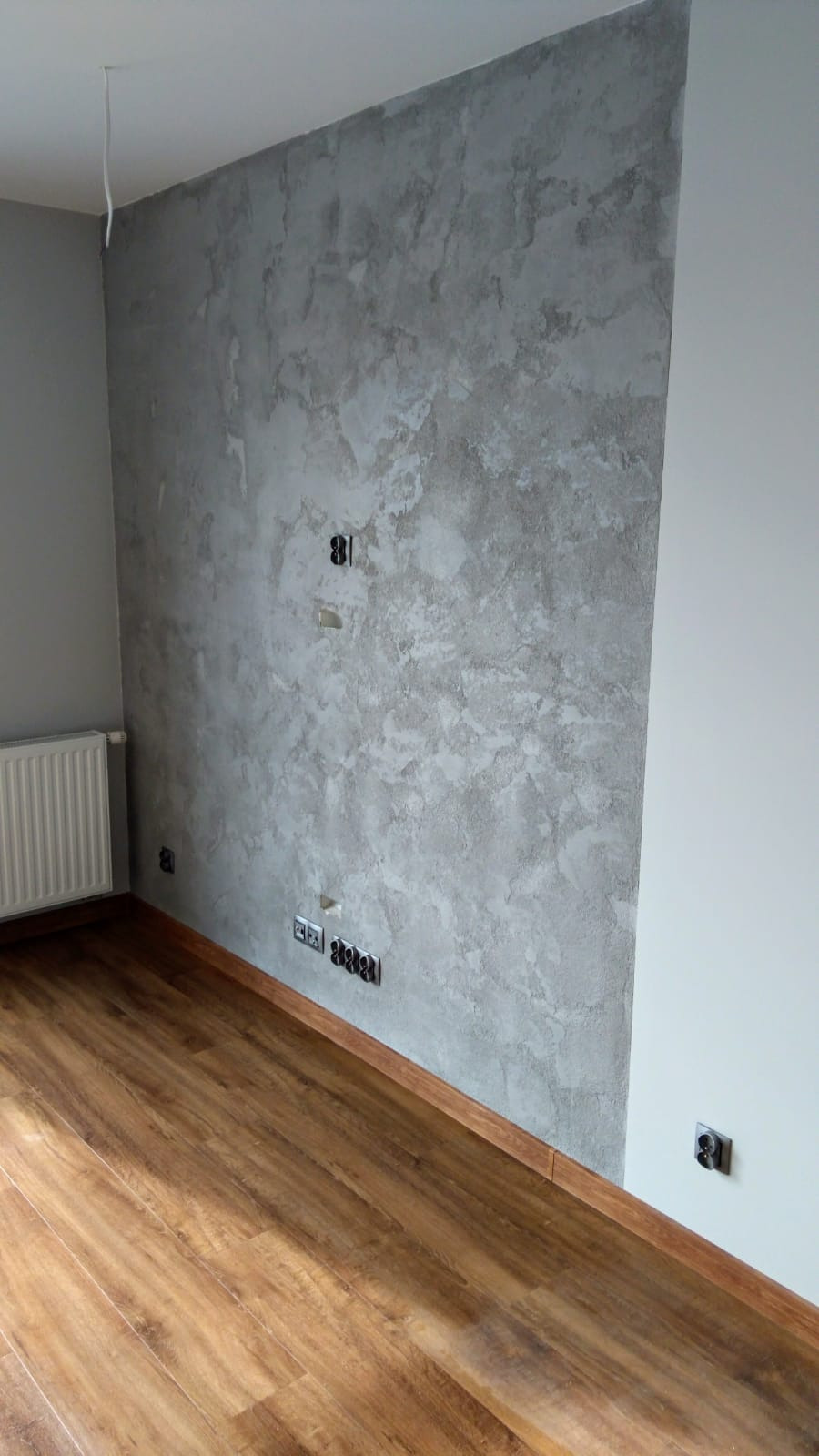 Pokój po remoncie: dekoracyjna szara ściana z efektem betonu, brązowe panele na podłodze, widoczne gniazdka elektryczne i wystający kabel ze ściany.