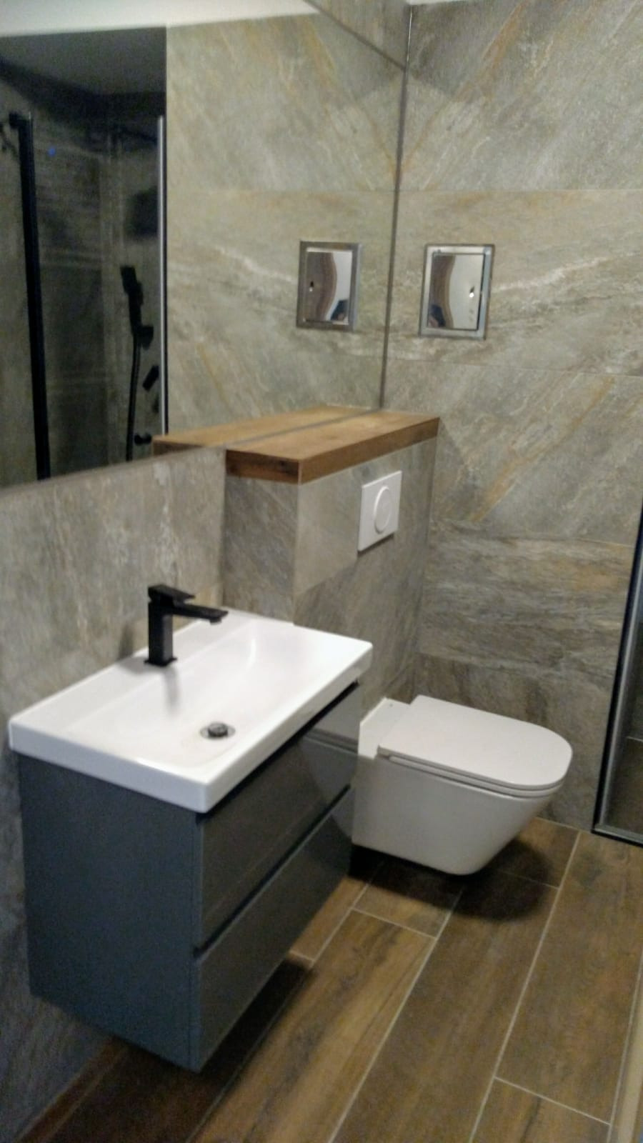 Nowoczesna łazienka z umywalką nablatową, szarą szafką pod umywalką, wiszącą toaletą i podłogą imitującą drewno, wykończona płytkami w odcieniach szarości i beżu.