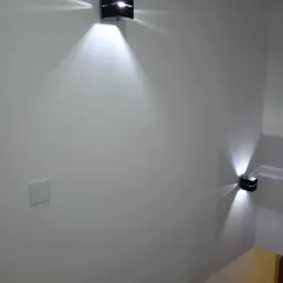 Dwa czarne, sześcienne kinkiety LED zamontowane na gładkiej, białej ścianie, emitujące promienie światła w górę i w dół, widoczny fragment żółtej ściany w rogu.