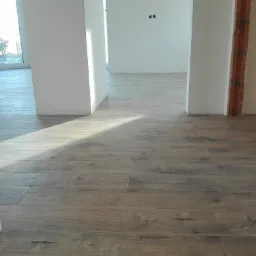 Ułożone podłużne płytki podłogowe imitujące drewno w jasnym, przestronnym wnętrzu w trakcie wykańczania, z widoczną instalacją w ścianie i promieniami słońca padającymi na podłogę.