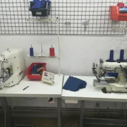 Dwie maszyny do szycia marki Texi na białych stołach, z widocznymi nićmi, nożykiem i stertą niebieskiego materiału, na tle ściany z siatką i pojemnikami na akcesoria.