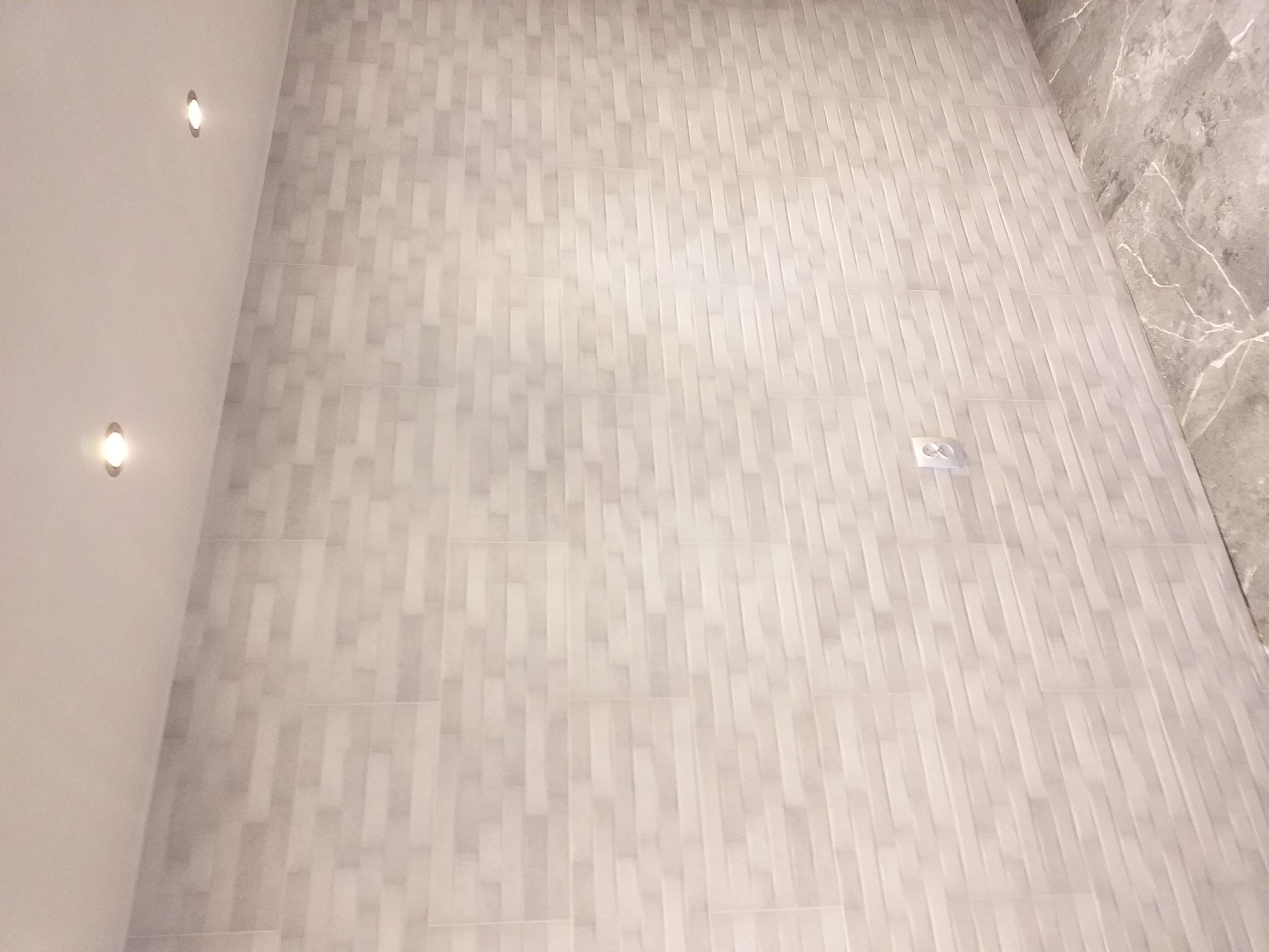 Jasnoszara ściana wyłożona pionowymi, prostokątnymi płytkami imitującymi kamień, z widocznym gniazdkiem elektrycznym i oświetleniem punktowym.