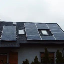 Panele słoneczne zamontowane na dachu domu jednorodzinnego z oknami dachowymi, widok z dołu na szarym tle nieba.