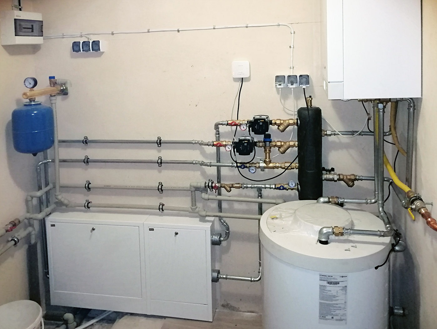 Kompletna instalacja grzewcza z piecem gazowym, zasobnikiem ciepłej wody i rozdzielaczem, widoczne liczniki ciepła i manometry, rury prowadzone wzdłuż ściany.