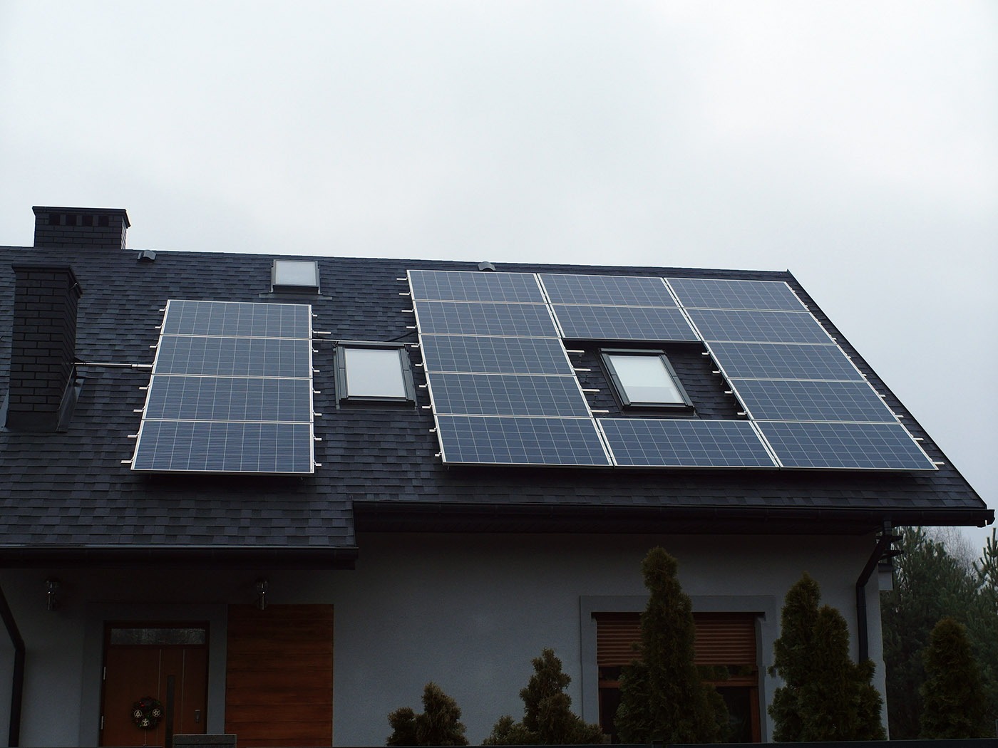Panele słoneczne zamontowane na dachu domu jednorodzinnego z oknami dachowymi, widok z dołu na szarym tle nieba.