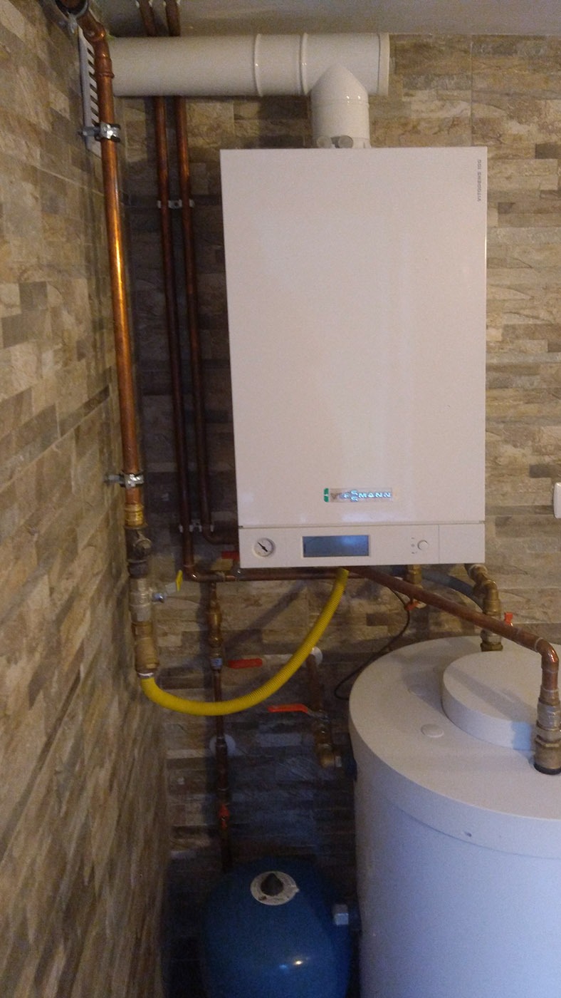 Biały piec gazowy Viessmann zamontowany na ścianie z imitacji kamienia, widoczne miedziane rury, żółty przewód gazowy i niebieski zbiornik wyrównawczy.