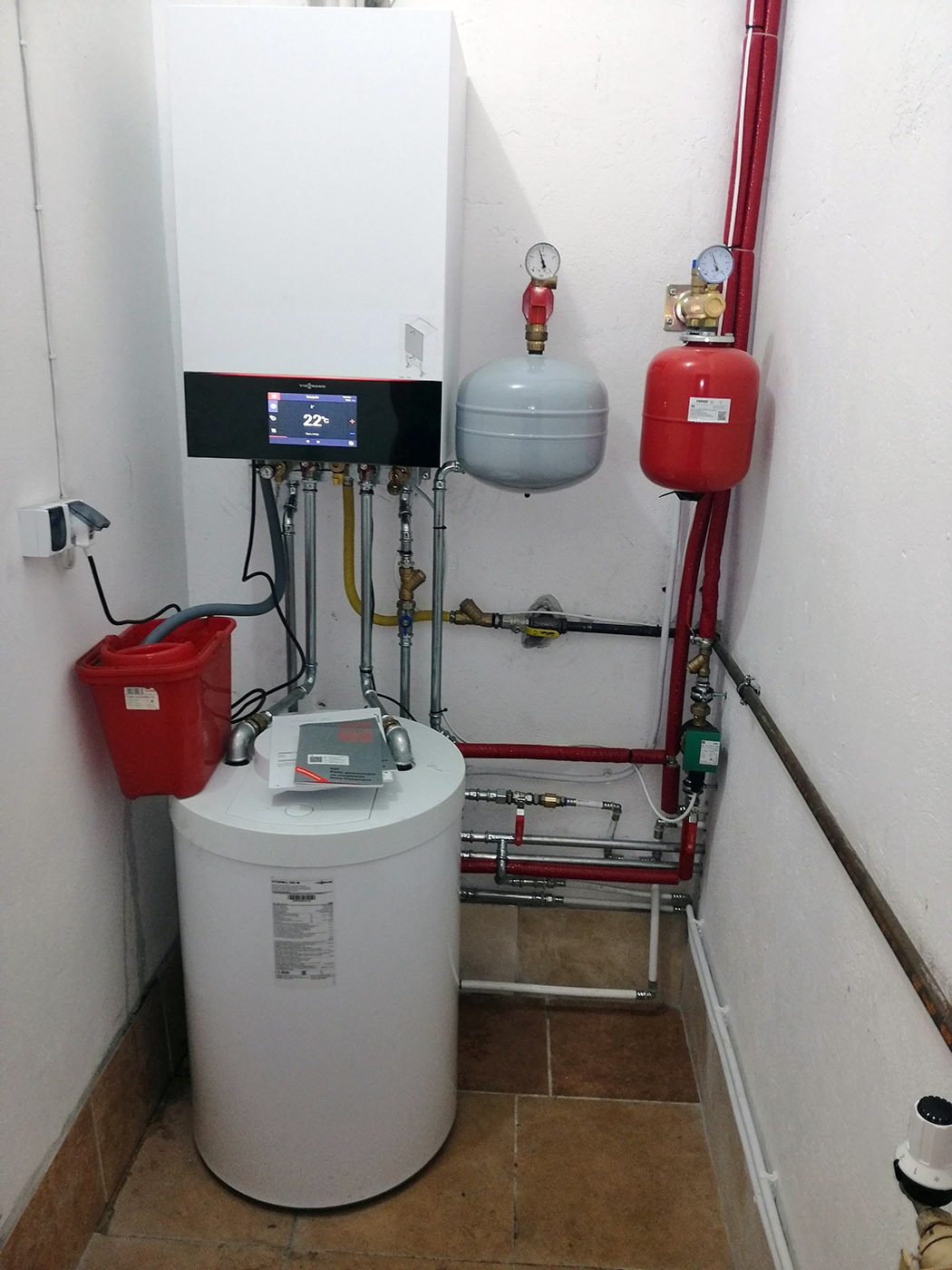 Nowoczesny, biały piec gazowy Viessmann z elektronicznym wyświetlaczem temperatury (22°C), podłączony do instalacji z czerwonymi rurami i zbiornikiem, zamontowany w kotłowni wyłożonej brązowymi...