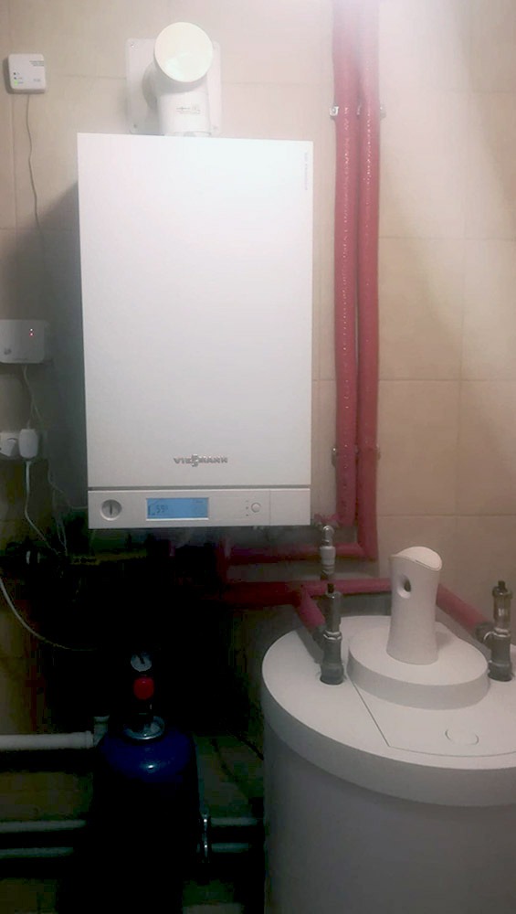 Biały piec gazowy Viessmann z wyświetlaczem, podłączony do instalacji z czerwonymi rurami, obok biały zbiornik na wodę i niebieska butla, widok z dołu.