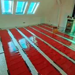 Montaż folii grzewczej pod ogrzewanie podłogowe w pomieszczeniu z oknami dachowymi i piecem kaflowym w rogu.