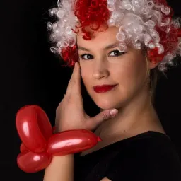 Portret kobiety w peruce z czerwono-białych loków, z czerwonymi ustami i balonowym motylem na nadgarstku, na czarnym tle.