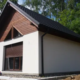 Niewielki, parterowy budynek z drewnianą g&oacute;rną częścią szczytu i czarną dach&oacute;wką, okno z roletą zewnętrzną, zabawka-traktor na tle, w otoczeniu zieleni.