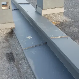 Szare kominy wentylacyjne z metalowymi obróbkami na dachu pokrytym papą, widok z góry.