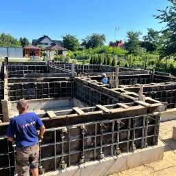 Budowa fundamentów: szalunek zbrojony, dwóch pracowników przy pracy, widok na działkę z domami w tle, słoneczny dzień.