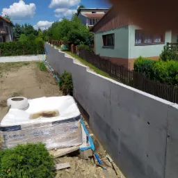 Świeżo wylany betonowy mur oporowy wzdłuż granicy działki, widoczne rury drenażowe i przygotowane podłoże pod instalację wanny spa HotSpring, domy mieszkalne w tle.