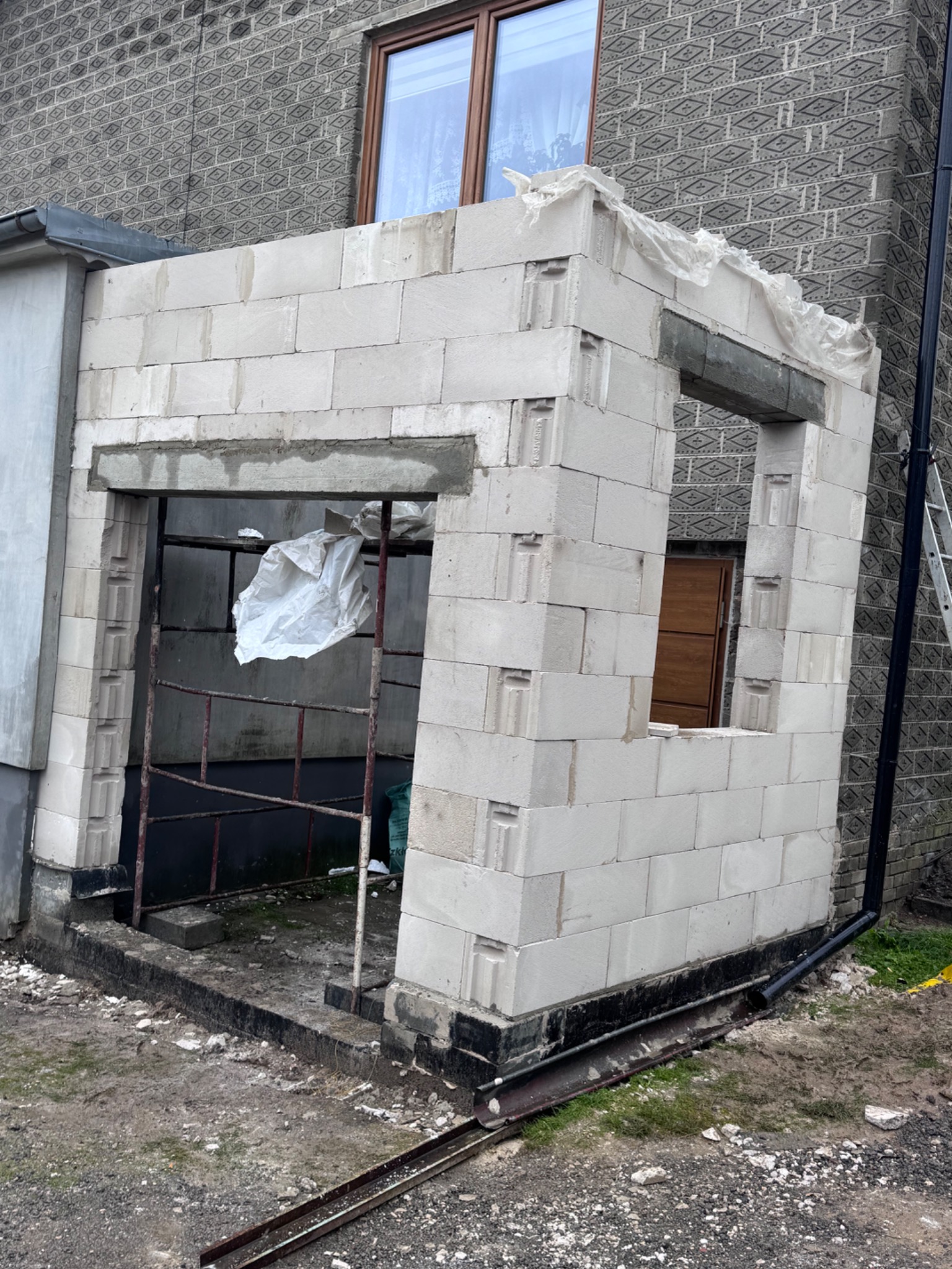 Budowa ścian parterowego budynku z bloczków betonowych, z widocznymi otworami okiennymi i drzwiowymi. W tle ściana budynku z oknem i rynna spustowa.