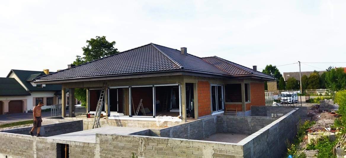 Budowa domu jednorodzinnego: fundamenty, ściany z czerwonej cegły, grafitowy dach. Widoczne okna przesuwne i drabina. Mężczyzna na fundamencie. Prace w toku.