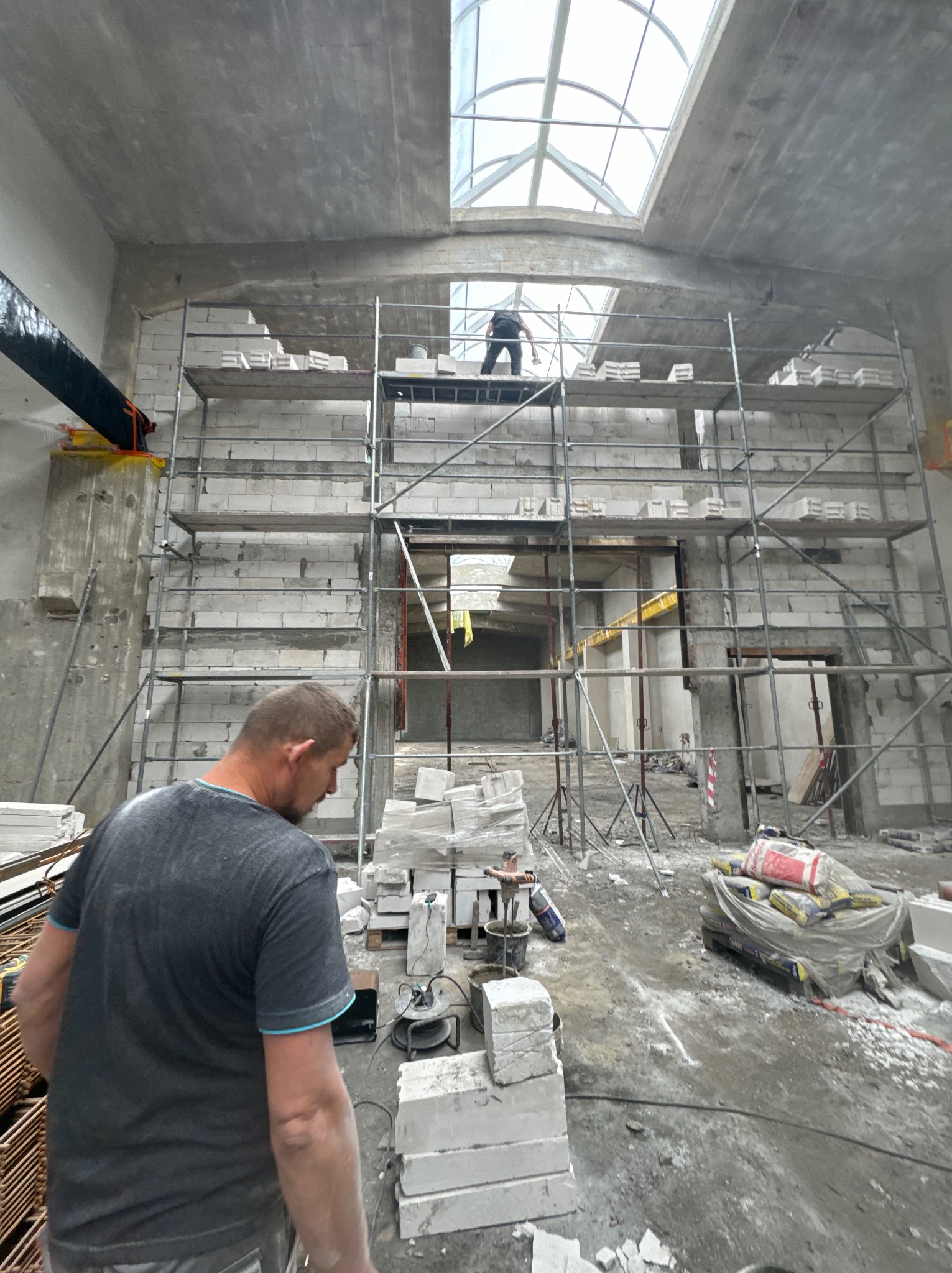 Wnętrze w trakcie murowania ścian z bloczków, widoczne rusztowania, narzędzia i materiały budowlane. W tle osoba na rusztowaniu i okno dachowe.