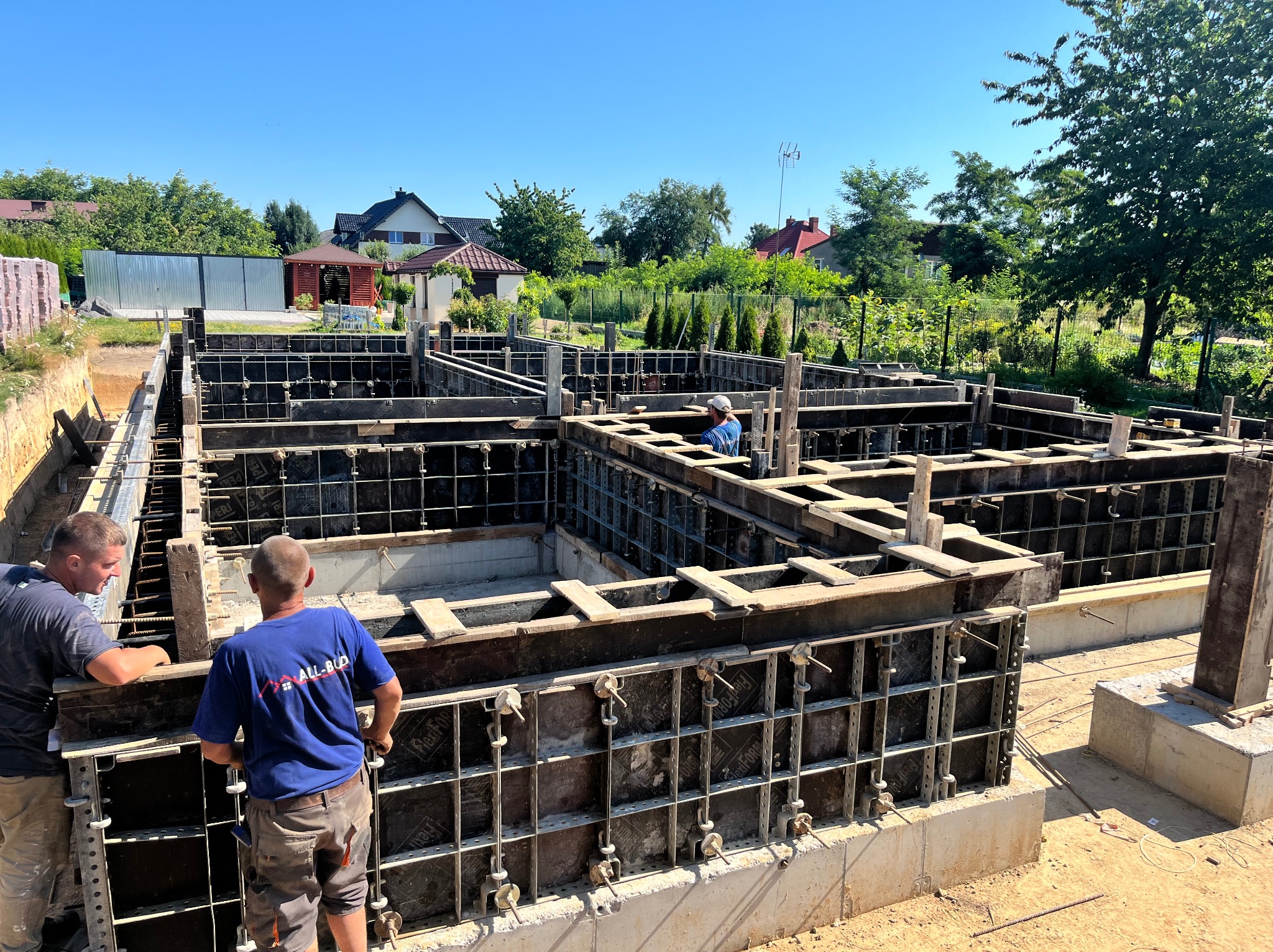 Budowa fundamentów: szalunek zbrojony, dwóch pracowników przy pracy, widok na działkę z domami w tle, słoneczny dzień.