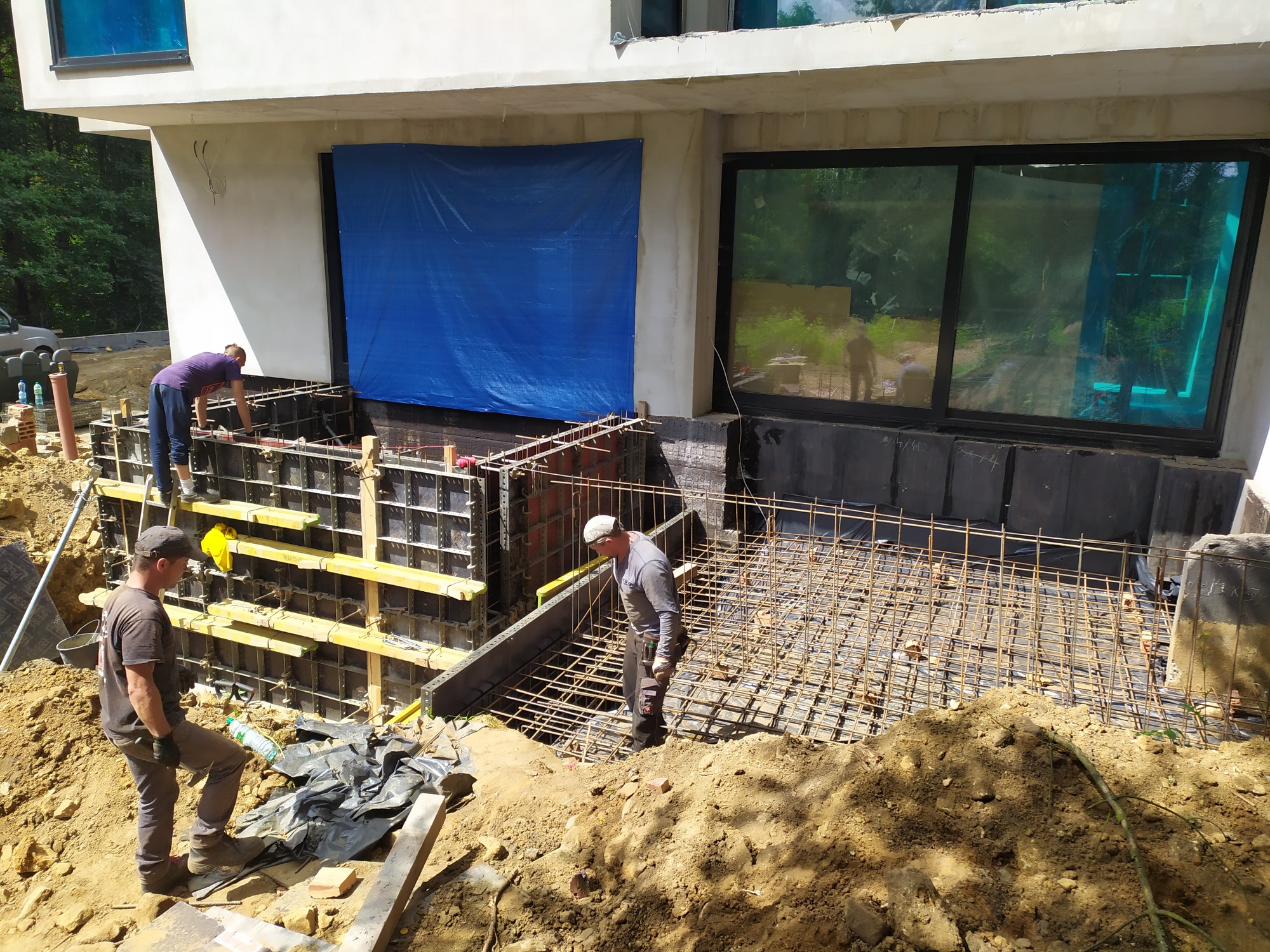 Trzech robotników pracuje przy szalunku i zbrojeniu fundamentów budynku w trakcie budowy, widoczne deski szalunkowe, stalowe pręty zbrojeniowe, wykop i elementy konstrukcyjne budynku.