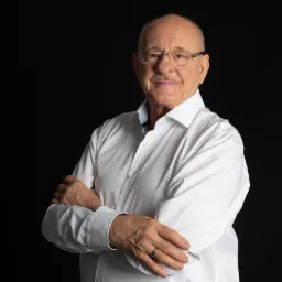 Portret starszego mężczyzny w okularach i białej koszuli, z założonymi rękami na czarnym tle.