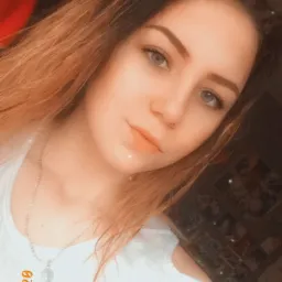 Portret młodej kobiety z długimi, ombre włosami i srebrnym naszyjnikiem, pozującej na tle ciemnego pokoju.