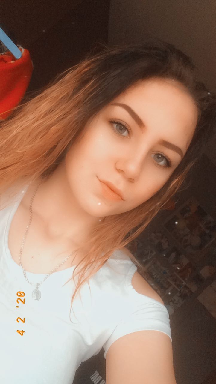 Portret młodej kobiety z długimi, ombre włosami i srebrnym naszyjnikiem, pozującej na tle ciemnego pokoju.