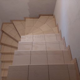 Usługi Remontowo Wykończeniowe Grotnik Mariusz - Wykończone schody z beżowych płytek ceramicznych z ryflowanymi krawędziami, widoczne z góry, ściana pomalowana na biało z widoczną strukturą.