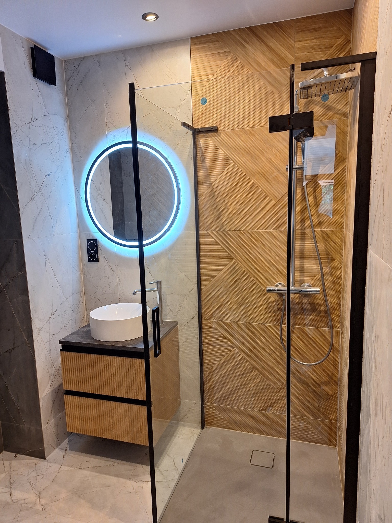 Nowoczesna łazienka z kabiną prysznicową typu walk-in z czarnymi profilami, umywalką nablatową z szafką z lamelami i okrągłym lustrem z podświetleniem LED.