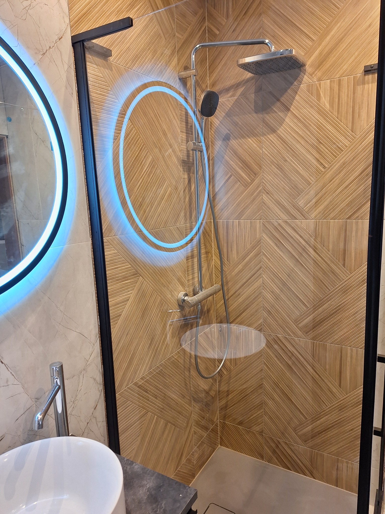 Nowoczesna kabina prysznicowa z deszczownicą i baterią, wykończona płytkami imitującymi drewno ułożonymi w geometryczny wzór. Umywalka ceramiczna i lustro z podświetleniem LED.