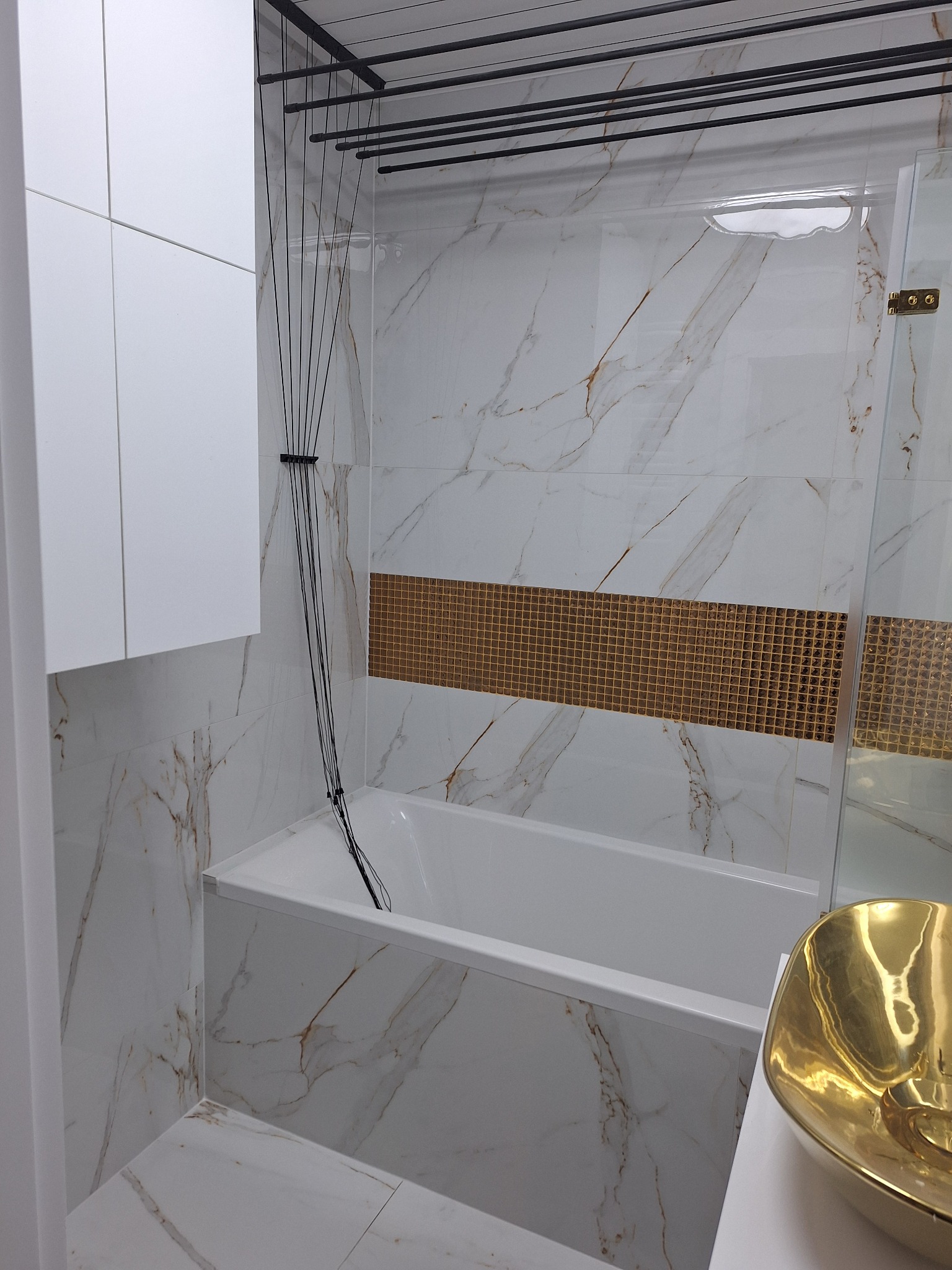 Nowoczesna łazienka z wanną obudowaną płytkami imitującymi marmur, złotym paskiem dekoracyjnym z mozaiki, złotą umywalką i suszarką sznurkową pod sufitem.