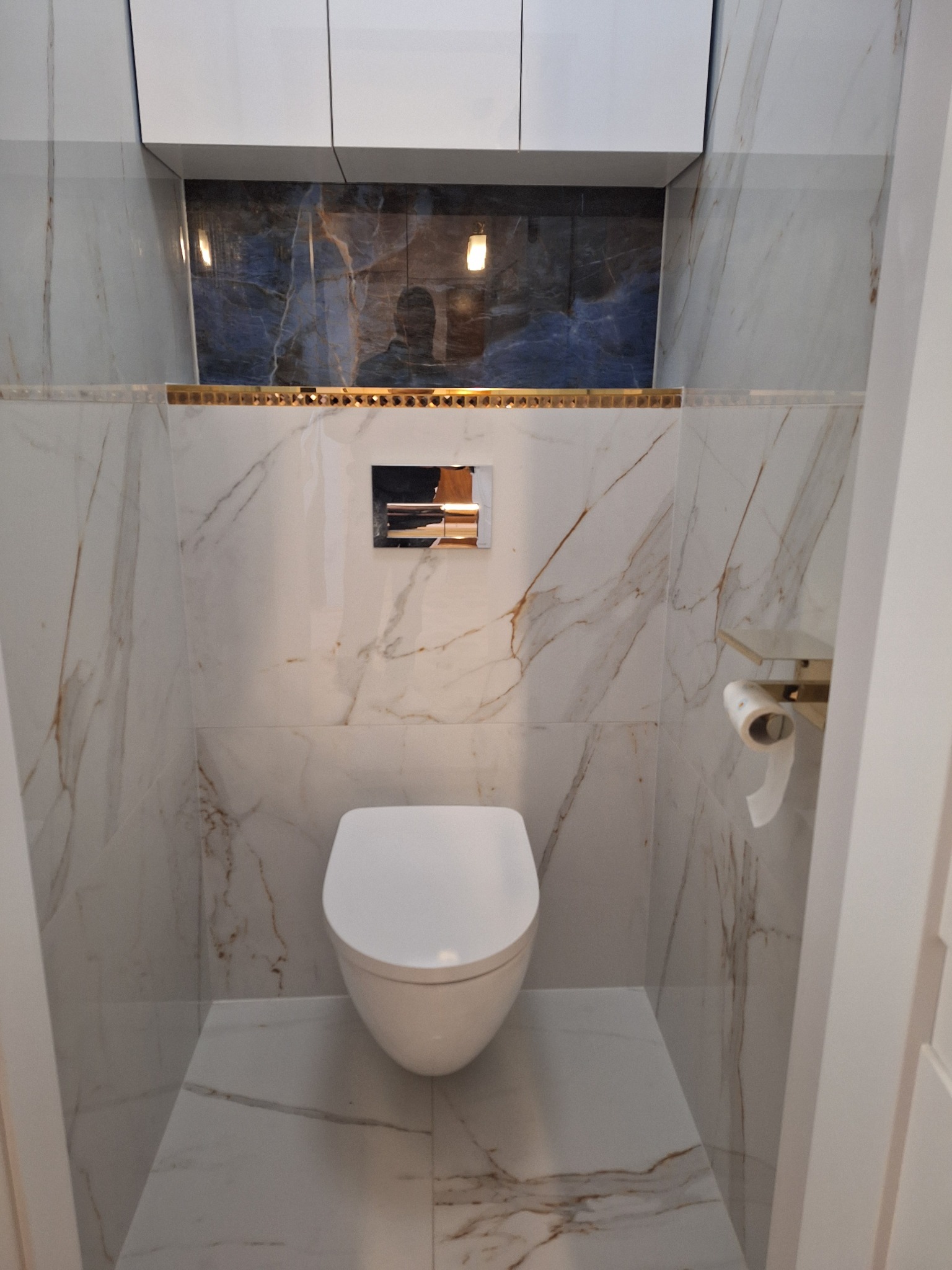 Nowoczesna toaleta podwieszana w łazience wykończonej białym marmurem z złotymi akcentami, widoczna zabudowa nad toaletą z niebieskim dekorem.
