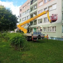 Żółty podnośnik koszowy na tle bloku mieszkalnego podczas pracy przy elewacji, widoczny pracownik w koszu na wysokości okna.