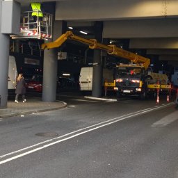 BEST LIFT Marcin Leśniewski - Pracownik w żółtej kamizelce w koszu podnośnika na wysięgniku, prawdopodobnie naprawia lub czyści elementy konstrukcji pod zadaszeniem parkingu miejskiego, na pierwszym planie ulica z liniami,...