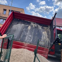 BEST LIFT Marcin Leśniewski - Czerwona wywrotka z otwartą skrzynią ładunkową, wypełniona piaskiem, widziana zza zielonego ogrodzenia posesji, na tle budynku mieszkalnego i błękitnego nieba z chmurami.