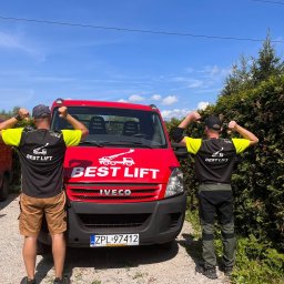 BEST LIFT Marcin Leśniewski - Dwóch pracowników firmy z Police, w koszulkach z logo firmy, prezentuje czerwony samochód dostawczy z logo firmy Best Lift na tle błękitnego nieba.