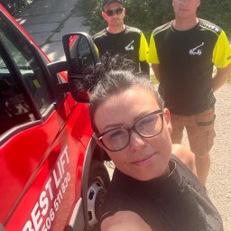 BEST LIFT Marcin Leśniewski - Autoportret kobiety w okularach na tle czerwonego samochodu z napisem 'BEST LIFT' i numerem telefonu, w tle dwóch mężczyzn w firmowych koszulkach z logo dźwigu