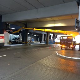 BEST LIFT Marcin Leśniewski - Żółty podnośnik koszowy z wysięgnikiem pod betonowym wiaduktem, pracownik w koszu, w tle zaparkowane samochody dostawcze.