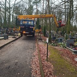 BEST LIFT Marcin Leśniewski - Żółty podnośnik koszowy na cmentarzu, z pracownikiem w koszu na tle drzew i nagrobków, liście na ziemi, pochmurny dzień.