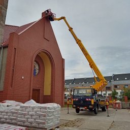 BEST LIFT Marcin Leśniewski - Żółty podnośnik koszowy przy ceglanym kościele z czerwoną dachówką, robotnik w koszu na wysokości, palety z zaprawą murarską na pierwszym planie, szare niebo w tle.