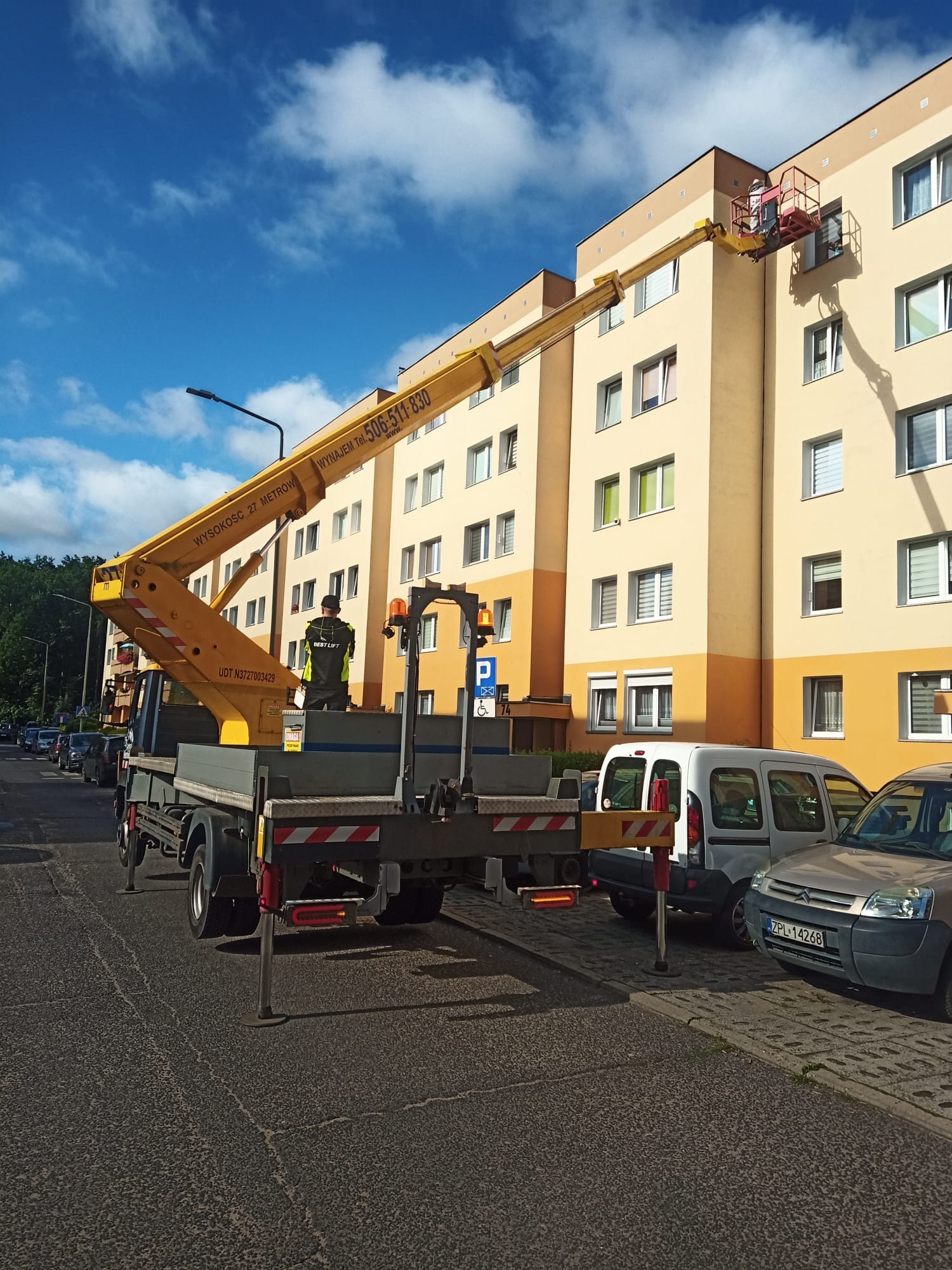 Żółty podnośnik koszowy na ulicy z operatorem w koszu przy ocieplonym bloku mieszkalnym, widok z tyłu platformy z podporami, zaparkowane samochody obok.