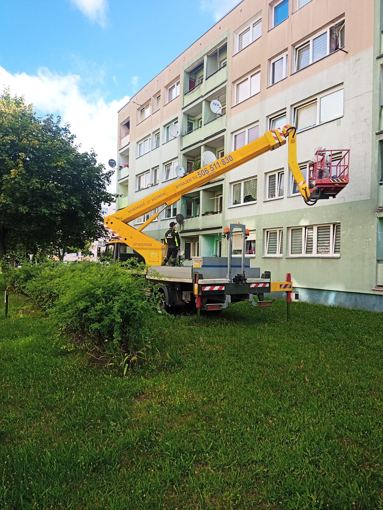 Żółty podnośnik koszowy na tle bloku mieszkalnego podczas pracy przy elewacji, widoczny pracownik w koszu na wysokości okna.