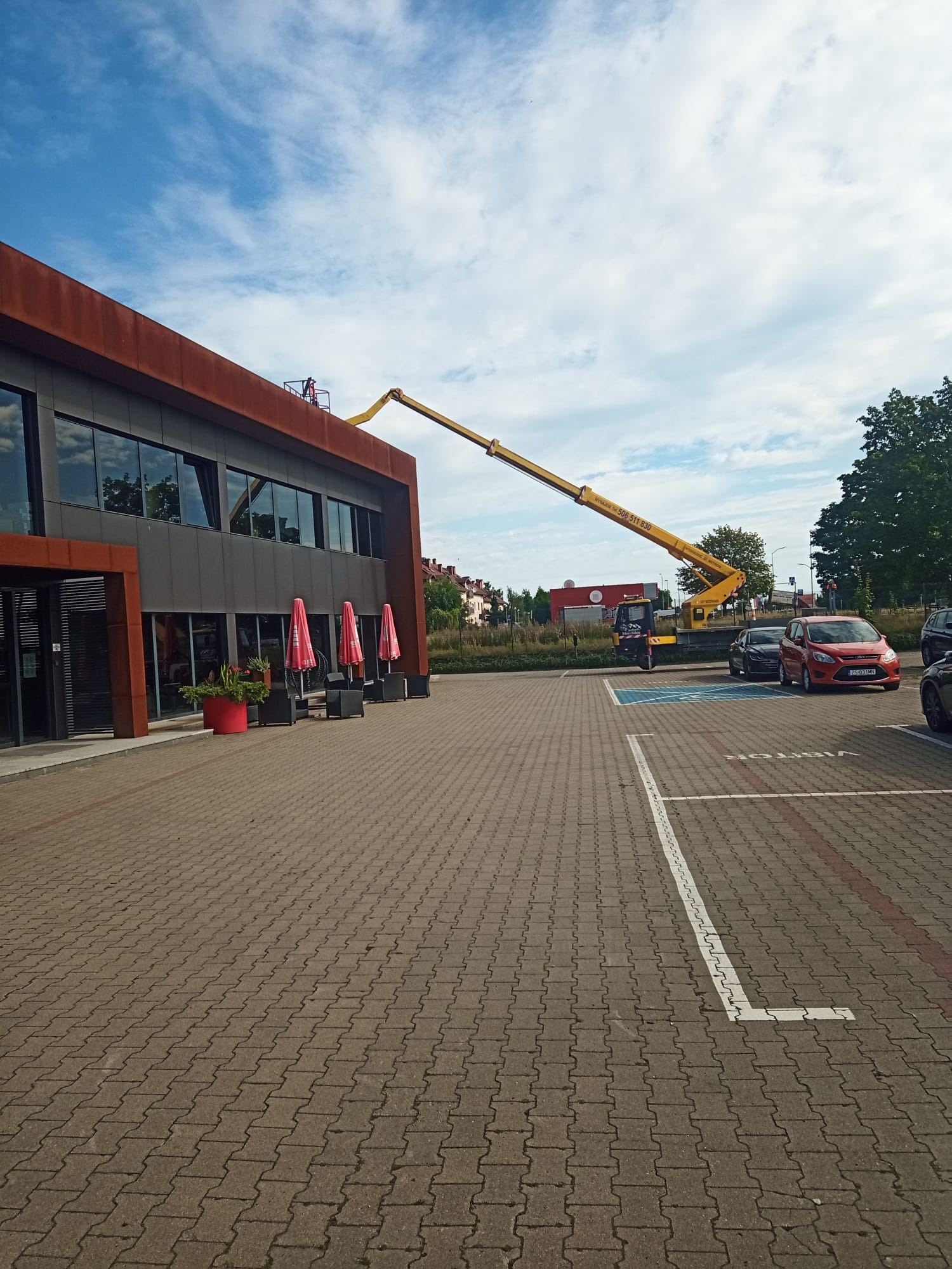 Żółty podnośnik koszowy z operatorem przy budynku biurowym z ciemną elewacją, widoczne zaparkowane samochody i błękitne niebo z chmurami.
